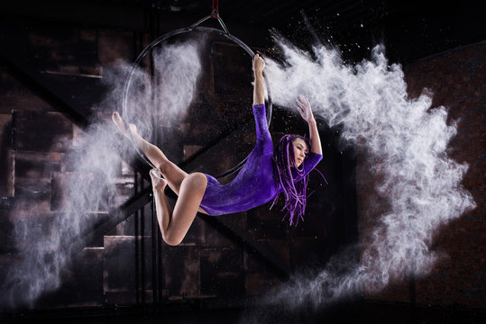 A young, slender girl in a lavender leotard and lilac dreads, doing gymnastics on the air ring, the Hoop and throws the hands of the white powder in the form of a cloud.