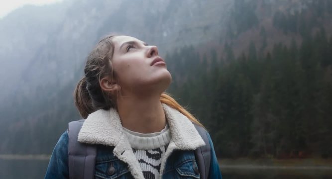 CU TO MED CRANE SHOT Portrait of beautiful Caucasian female hiker with backpack standing near beautiful lake in Alps, enjoys the rain. 4K UHD 60 FPS SLO MO