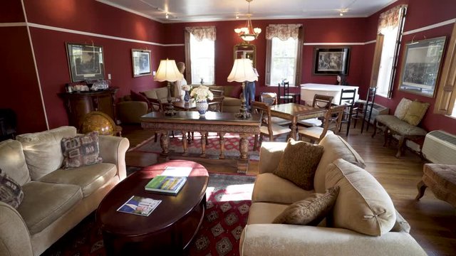 A woman walks into the room from camera left while an elderly man stands and looks around in a large lounge filled with couches and seating and a grand piano.