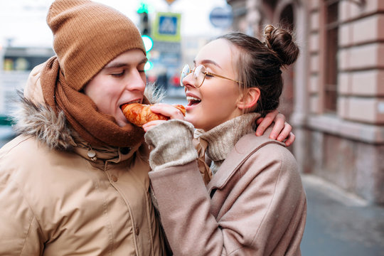 Beautiful couple in love winter street portrait. Pretty girl feeding her boyfriend with croissant
