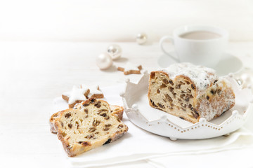 criststollen, typical german christmas cake with raisins and fruits, next to a cup of coffee, cinnamon stars and baubles on a white wooden background with copy space