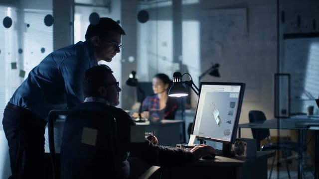 Late at Night Two Industrial Engineers Discuss 3D Component Design Made with CAD Program on Personal Computer. Office Looks Modern with Lots of Blueprints on the Walls.