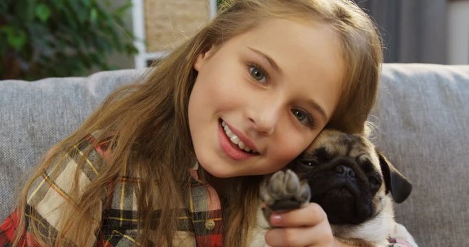 Close up of a cute blonde girl hugging a pug and waving its paw on the couch at home. Inside. Portrait