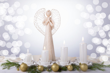 A white angel and four white Advent candles, Christmas tree branches are decorated with golden balls. The imminent background of Advent and Christmas
