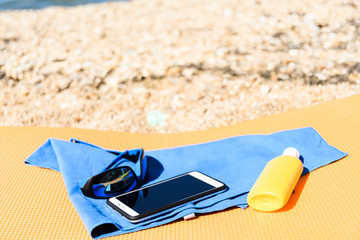 Sunglasses lying next to a smartphone on a microfiber towel next to the sunscreen on the travel rug on the beach