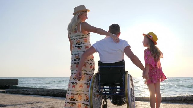 invalid husband in wheelchair hug pregnant wife with big belly and child close-up on background sea in backlight, portrait of happy family