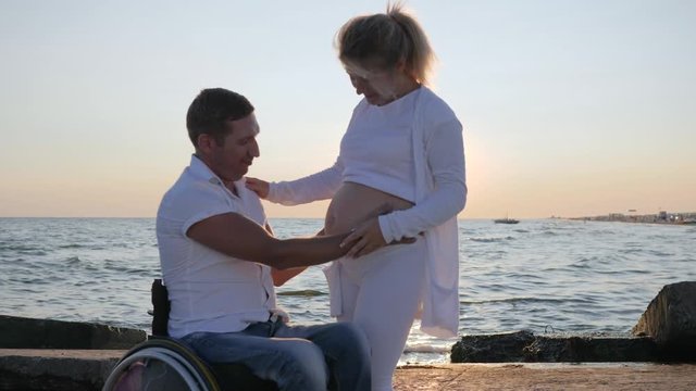 lovers disabled man on wheelchair and pregnant woman, family in anticipation of child at sunset background sea in the backlight