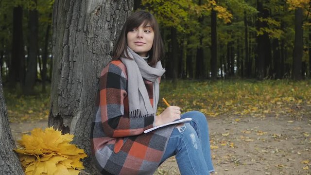 Wonderful autumnal view. Charming young happy smiling woman sitting on the grass in a park, take notes in notebook.