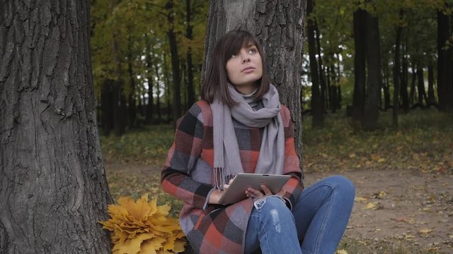 Wonderful autumnal view. Charming young think woman sitting on the grass in a park, use tablet