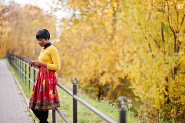 African american girl at yellow and red dress at golden autumn fall park with tablet at hands.