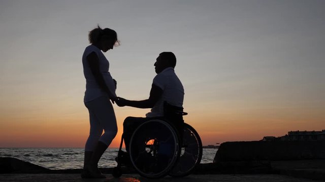silhouettes disabled husband in wheelchair holds arm wife with big belly, happy couple