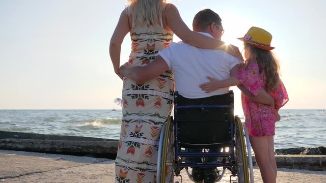 husband in wheelchair and wife with big belly hug close-up family looking at waves