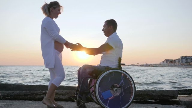 invalid spouse into wheelchair with pregnant female dancing at sunset, happy handicapped couple waiting for baby