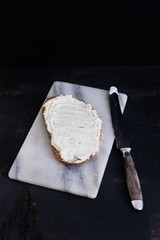 Slice of typical German sunflower seed wholegrain bread with herbed cream cheese spread on marble cutting board with vintage knife next to it on black wooden background
