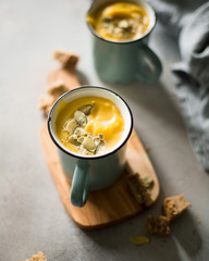 Pumpkin soup with coconut milk, pumpkin seeds and spices in vintage blue mugs on a gray concrete background. Atmospheric dark food photo. 