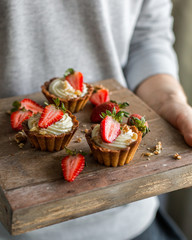Mini tartlets with cheese cream and fresh strawberries, sprinkled with nuts on a wooden box. Food photo with hands.