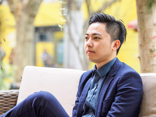 Portrait of a handsome Chinese young man in blue suit sitting outside and looking at camera.