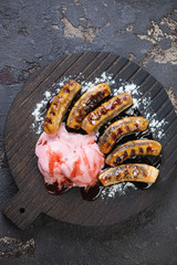 Black wooden serving board with roasted mini bananas, ice cream, sauce and powdered sugar on a brown stone surface, flat-lay