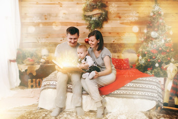 Young happy parents with little child son reading fairy tale in book near Christmas tree in decorated New Year wooden room at home. Lifestyle, family and holiday 2018 concept. Magical highlight effect