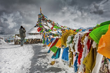 Photographer taking photos of Tibetan prayer flags on a snow mountain - obrazy, fototapety, plakaty