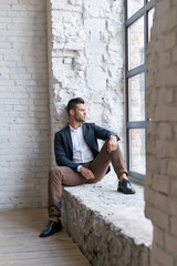 Man in dark blazer, aqua shirt and brown trousers at white brick wall and dark window