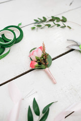 Scissors, ribbons and greenery lie around wedding boutonniere on white table