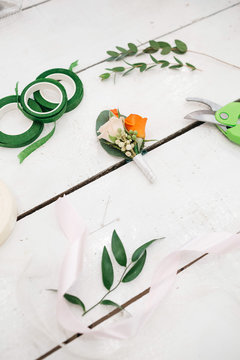 Scissors, ribbons and greenery lie around wedding boutonniere on white table