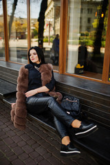 Fototapeta premium Fashion outdoor photo of gorgeous sensual woman with dark hair in elegant clothes and luxurious sleeveless fur coat and with backpack sitting on bench at autumn city.