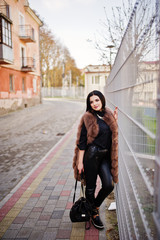Naklejka premium Fashion outdoor photo of gorgeous sensual woman with dark hair in elegant clothes and luxurious sleeveless fur coat against iron fence at autumn city.