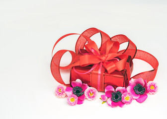 Festive gift box with a bow of red color. Lies on a white background. Next to pink flowers.