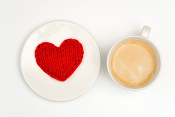 Fototapeta na wymiar A cup of coffee is on a white table. There is a knitted red heart on the plate.