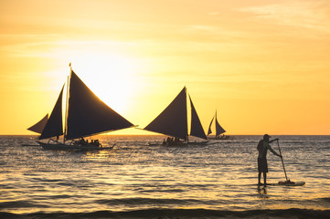 Silhouette of sailing boats at sunset in Boracay island - Unrecognizable man with paddle surfing - Exclusive travel destination in Philippines - Warm vintage filtered look - Focus in foreground