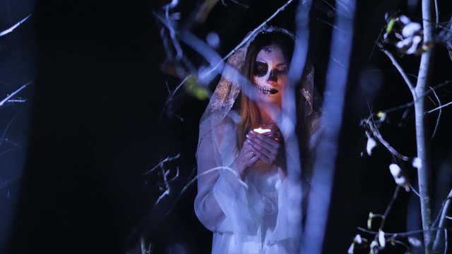 The young woman with skeleton make-up for Halloween in the white bride dress holding in the arms a burning wax candle in the dark forest at night. 4K