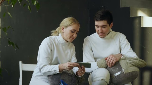 Two young fencers man and woman watching fencing tutorial on smartphone and sharing experience after training indoors