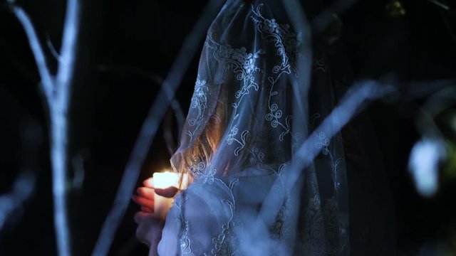 The young woman with skeleton make-up for Halloween in the white bride dress holding in the arms a burning wax candle and blowing it out in the dark forest at night. 4K