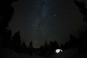 Fototapeta premium White tent under the starry sky and the milky way in the trees in the winter mountains at night