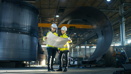 Male and Female Industrial Engineers Look at Project Blueprints While Standing Surround By Pipeline...