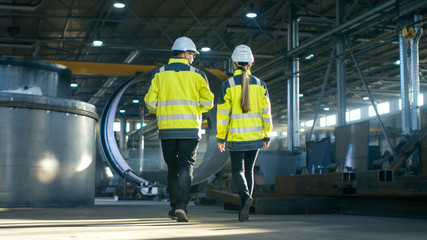 Back View Shot of Male and Female Industrial Engineers Having Discussion While Walking Through Heavy Industry Manufacturing Factory. Big Metalwork Constructions, Pipeline Elements Lying Around.