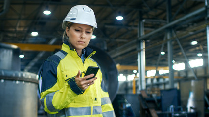 Female Industrial Worker in the Hard Hat Uses Mobile Phone While Walking Through Heavy Industry...
