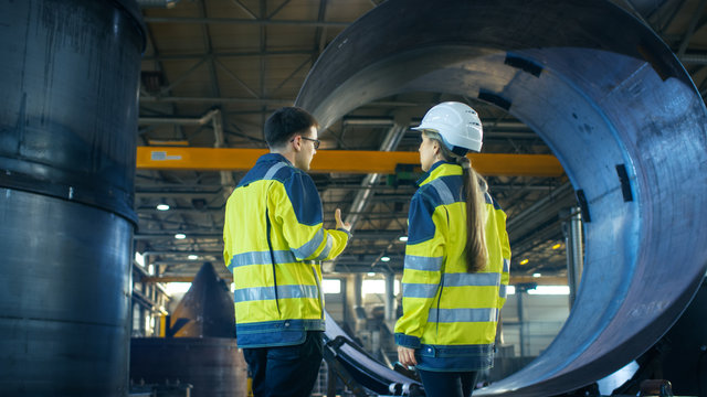 Male Industrial Worker and Female Chief Engineer in a Hard Hat Discuss Metalworking Factory's New Project. They Stand in the Middle of Heavy Industry Facility.
