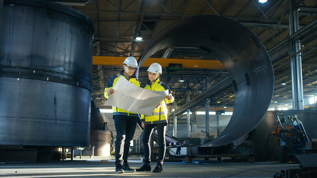 Male and Female Industrial Engineers Look at Project Blueprints While Standing Surround By Pipeline Parts in the Middle of Enormous Heavy Industry Manufacturing Factory