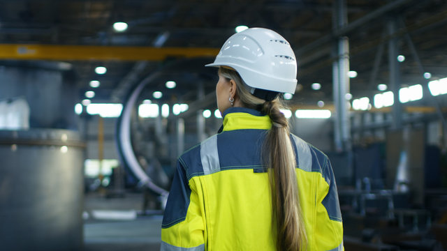 Shot of Female Industrial Worker in the Hard Hat Walking Through Heavy Industry Manufacturing Factory. In the Background Various Metalwork Project Parts Lying