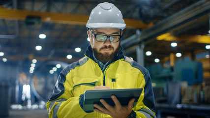 Industrial Engineer in Hard Hat Wearing Safety Jacket Uses Touchscreen Tablet Computer. He Works at the Heavy Industry Manufacturing Factory.
