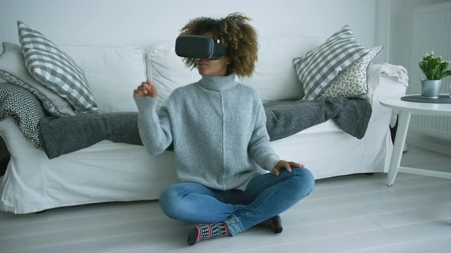 Excited young woman in casual clothing relaxing at home in VR glasses while sitting on floor of living room and touching air with hand.