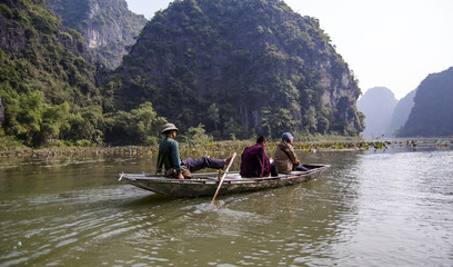 Rowers use their legs to move the oars. People on the boat are moving along the Ngo Dong River between the rocks. Hoa Lu Tam Coc, Hoi An Ancient Town, Vietnam.