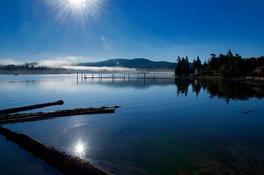 The morning sun rises over Sooke harbour burning off the fog casting its reflection into the calm waters.