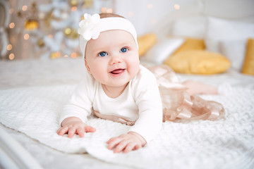 Baby girl wearing cute dress and headband, lies on a white cover in festively decorated room with garland of lights. With surprise watches in the camera, on a background a set of bright fires.