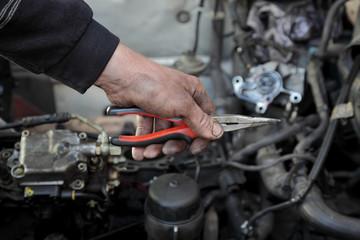 Dirty mechanic hand holding long nose pliers in hand with car engine in background