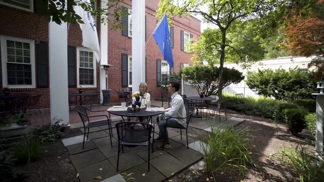 Camera orbits around a mature mother and hipster teen son sit at an outdoor table and wait for their meal to be delivered.