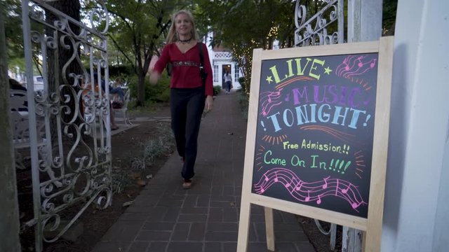 Beautiful blonde woman in yoga pants walks out of a garden with a sign for live music.
